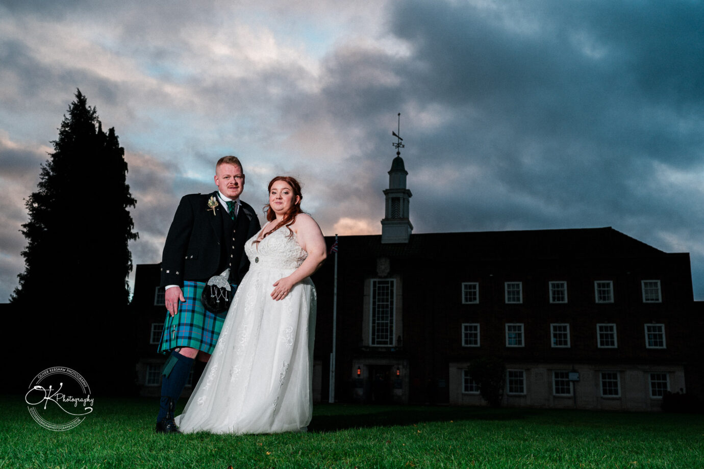 Bride and groom standing on grass in front of Derby Conference Centre, with a dramatic sky in the background.