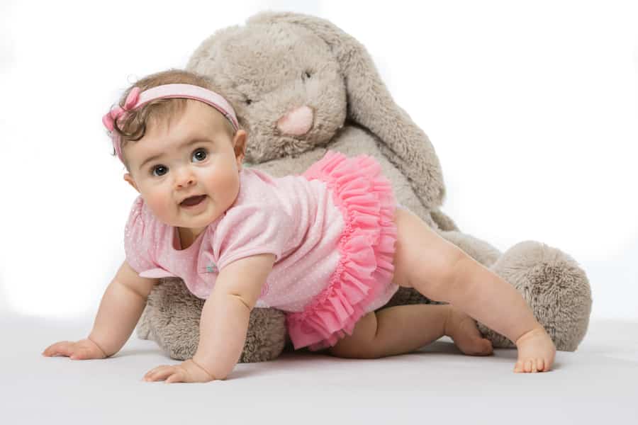 Baby in a pink outfit crawling in front of a large grey stuffed bunny.